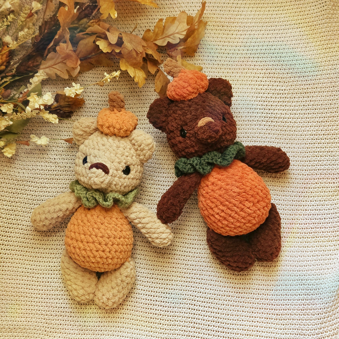 Two crocheted teddy bear plush toys styled as autumn pumpkins, one light beige and one dark brown, both with orange yarn bellies and hats and green yarn scarves, placed on a cream-colored textured blanket with dried leaves in the corner.