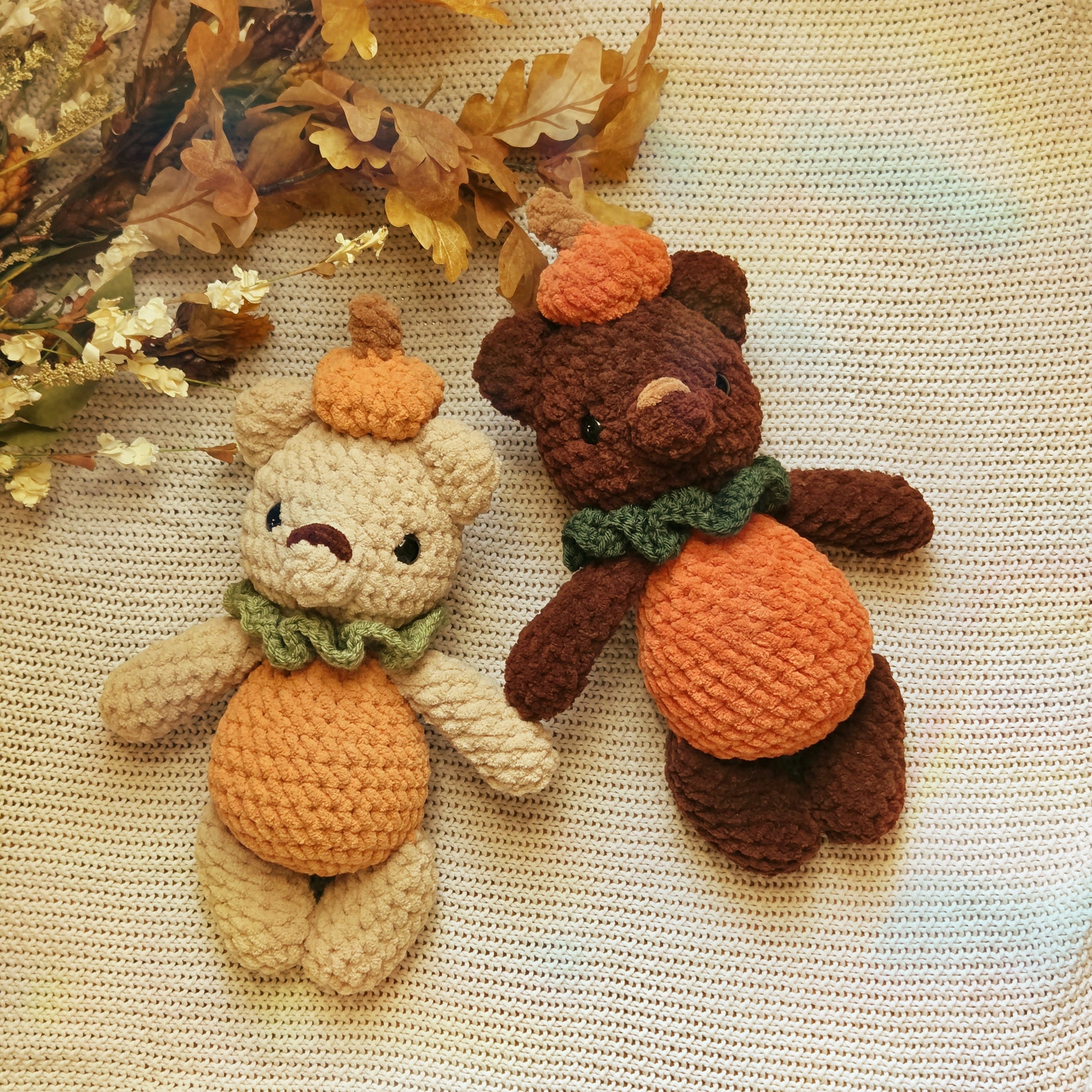Two crocheted teddy bear plush toys styled as autumn pumpkins, one light beige and one dark brown, both with orange yarn bellies and hats and green yarn scarves, placed on a cream-colored textured blanket with dried leaves in the corner.
