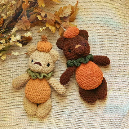Two crocheted teddy bear plush toys styled as autumn pumpkins, one light beige and one dark brown, both with orange yarn bellies and hats and green yarn scarves, placed on a cream-colored textured blanket with dried leaves in the corner.