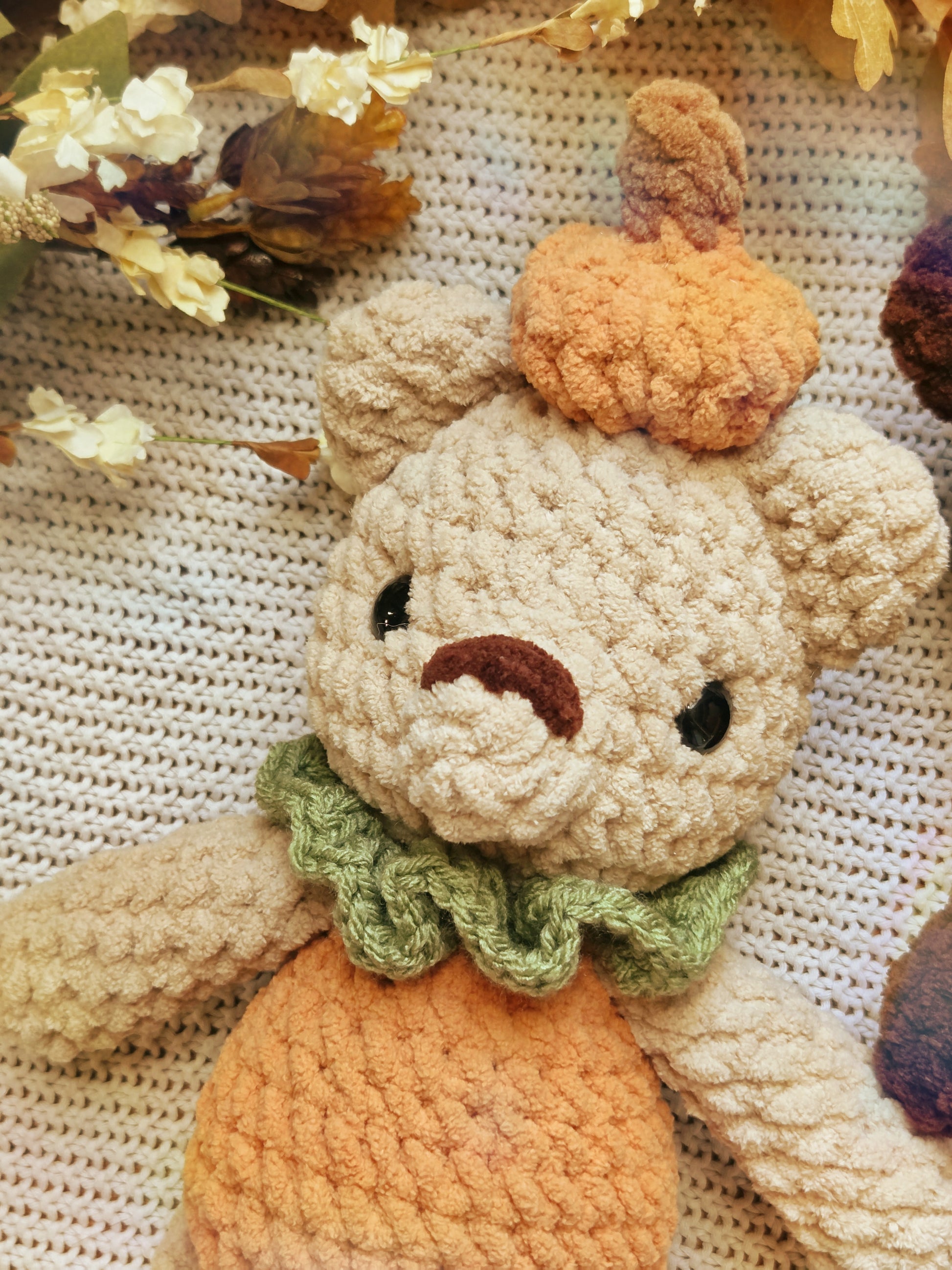 A close-up of a crocheted plush bear styled like a pumpkin, with a beige body, orange yarn stem and lower body, and a green ruffled collar, resting on a textured cream knit surface with dried flowers.