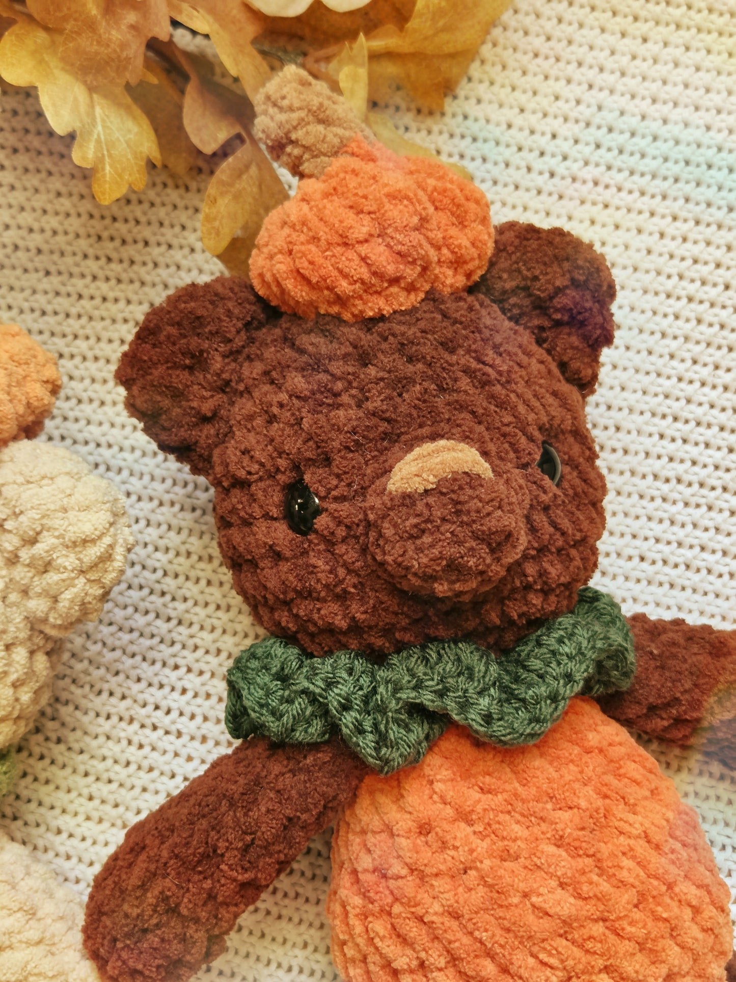 A close-up of a crocheted teddy bear dressed as an autumn pumpkin, featuring a brown body, orange yarn for a pumpkin-shaped hat and belly, and a green yarn scarf, resting on a textured cream knit blanket with dried leaves.