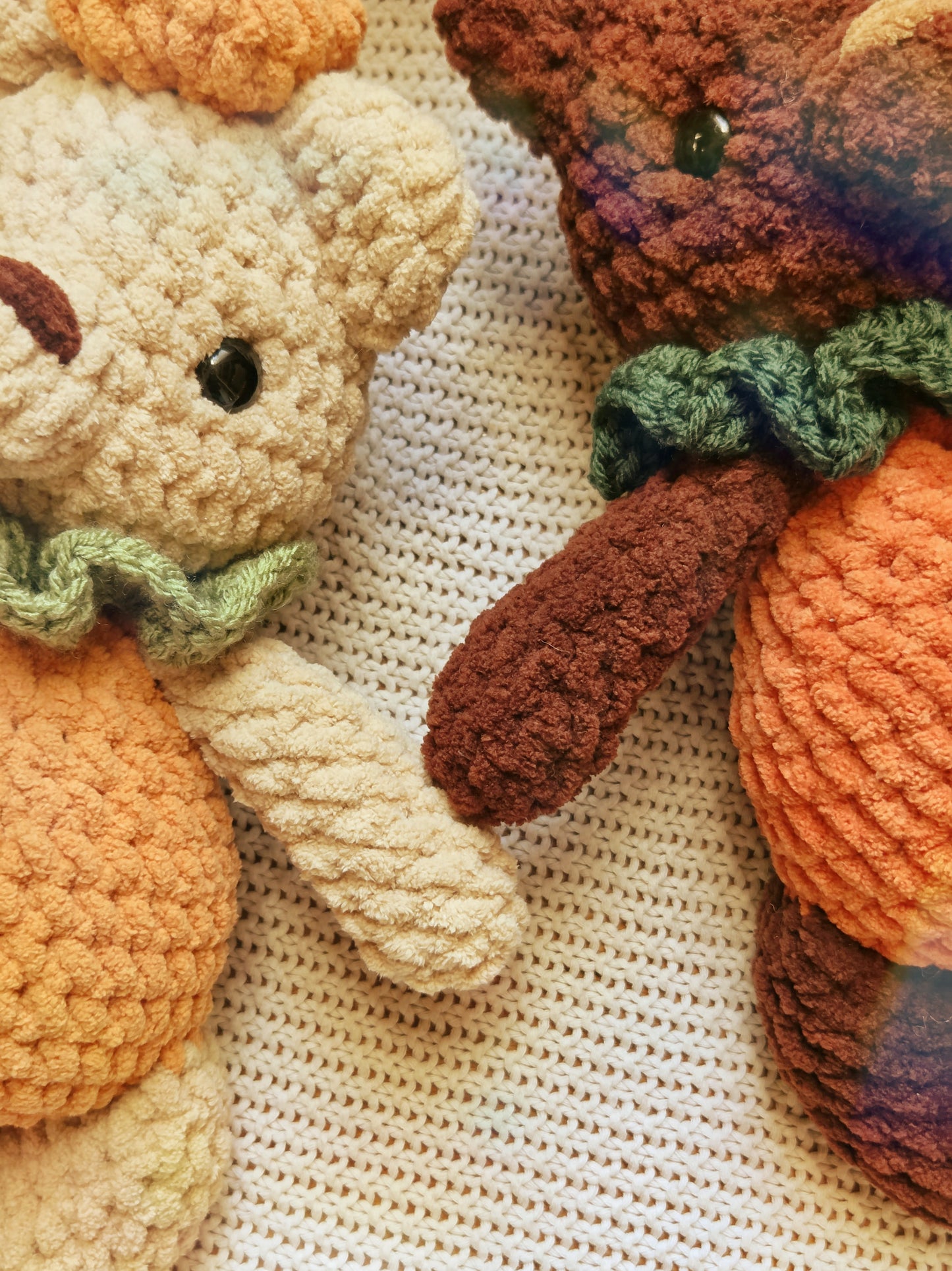 Close-up of two crocheted teddy bears on a cream knit blanket. One bear is cream with orange and green accents, and the other is dark brown with orange and green accents.