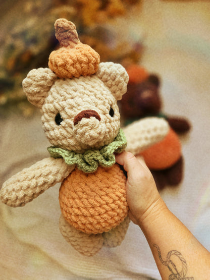 A handmade crocheted teddy bear plush toy styled like a pumpkin, held by a hand. The bear has a beige body, an orange pumpkin belly and hat, and a green ruffled collar.