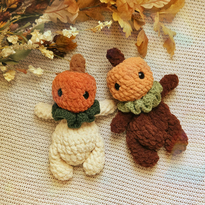 Two cute crocheted plush bear toys styled as pumpkins and acorns on a textured cream knit blanket with dried autumn leaves. One toy is cream-colored with an orange pumpkin head and dark green scarf.